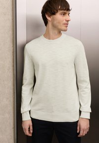 Jeune homme debout contre une porte d'ascenseur métallique, portant un pull beige clair et un pantalon foncé, regardant vers sa droite.