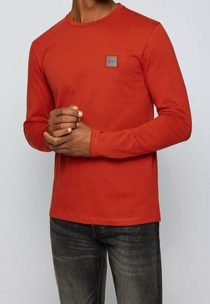 Homme portant une chemise à manches longues couleur rouille avec un petit patch logo "BOSS" sur la poitrine et un jean gris foncé, les mains croisées.