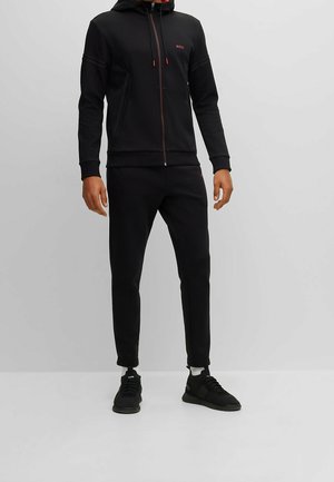 Mężczyzna ubrany w czarną bluzę z zamkiem i czarne spodnie dresowe oraz czarne trampki na jednolitym szarym tle.