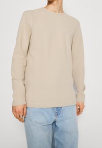 Beige långärmad skjorta i texturerad tyg, med rund halsringning och diskret logotypdetalj på bröstet, kombinerad med ljusblå jeans.