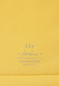 Žlutá tkanina má hladkou strukturu s prošívaným vzorem. Název značky "Joules" a "Adventure Awaits" jsou vytištěny světle modrou barvou.