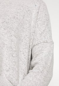 Světlý šedý pletený svetr s texturovaným povrchem a mírně zatočenými rukávy. Měkká, pohodlná látka s jemnými tmavšími vlákny po celé ploše.