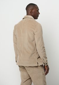 Beige corduroy jas met een kraag, zichtbare stiksels en zijsplitten. Heeft knoopsluiting bij de manchetten en een gestructureerd ribbelpatroon.
