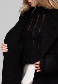 Mustat neuleet, joissa on läpikuultavia raidallisia yksityiskohtia, yhdistettynä mustaan takkiin. Mallilla on pitkät valkoiset kynnet ja pehmeät pinnat.