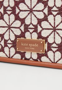 Borsa in tessuto texturizzato con un motivo floreale bianco su sfondo bordeaux, base in pelle marrone e una placca con logo dorato.