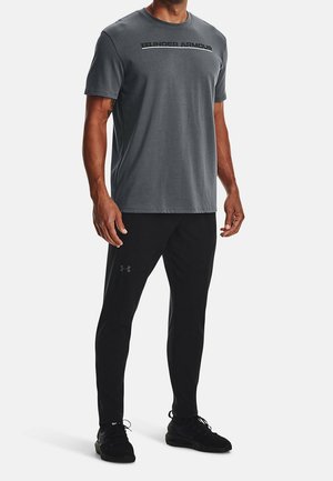 Camiseta atlética gris de manga corta con el logo de "Under Armour". Combinada con pantalones atléticos negros y zapatos negros, con un diseño elegante.