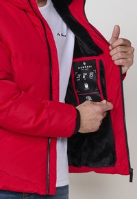 Chaqueta aislante roja con cremallera frontal, puños negros y un suave forro de piel sintética negra. La etiqueta interior muestra las instrucciones de cuidado y características.