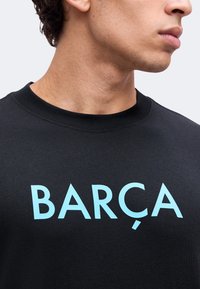 Νεαρός άνδρας φοράει μαύρο T-shirt με μεγάλα ανοιχτό μπλε γράμματα που σχηματίζουν το "BARÇA" στο στήθος, κοιτάζοντας δεξιά.
