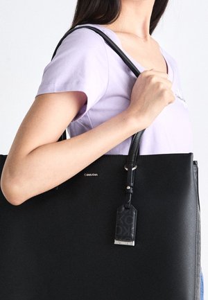 Osoba ubrana w jasnofioletową koszulę, trzymająca dużą czarną torbę na ramię Calvin Klein na tle jednolitego koloru.