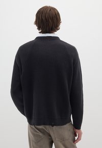 Schwarzer gerippter Strickpullover mit lockerer Passform, ausgestattet mit einem weiten Kragen und einer strukturierten Oberfläche, kombiniert mit hellgrauen Hosen.