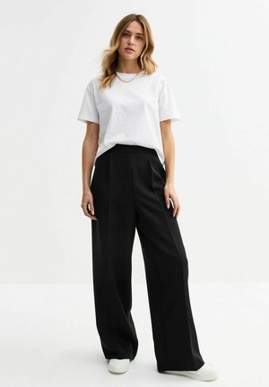 T-shirt bianco a maniche corte abbinato a pantaloni larghi neri. I pantaloni presentano un design drappeggiato e sono realizzati in un tessuto liscio.