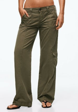 Femme portant un pantalon cargo vert olive avec des poches avant et latérales, debout les mains dans les poches et des sandales noires à bouts ouverts.