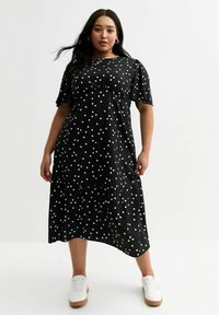 Schwarzes Kleid mit weißen Punkten, kurzen Puffärmeln und einem gestuften Saum. Getragen mit weißen Turnschuhen und aus einem glatten Stoff.
