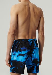 Mężczyzna bez koszulki widziany z tyłu, w niebieskich i czarnych kąpielówkach w tropikalny wzór z elastycznym paskiem i boczną kieszenią.