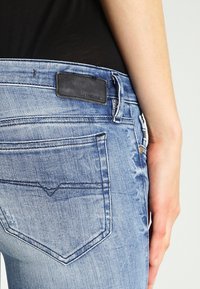 Jeans de mezclilla azul claro con un bolsillo trasero y etiqueta de cuero negro de la marca Diesel, llevados por una persona con una camiseta negra, mano visible en el costado.