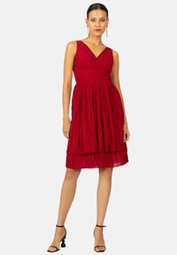 Robe plissée rouge avec un décolleté en V, un corsage ajusté et une jupe superposée. Présente des fines bretelles et une texture lisse, associée à des talons noirs à brides.