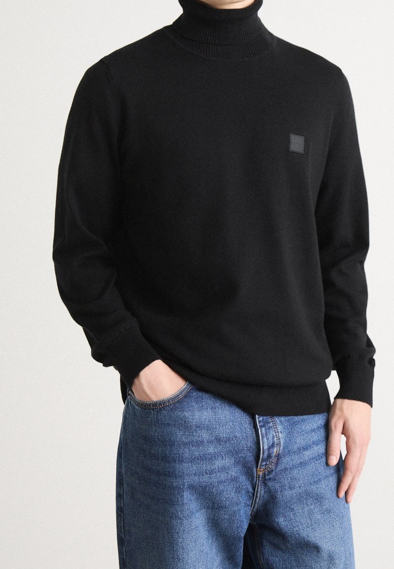 Pull à col roulé noir en matière lisse, avec une coupe ajustée, un col côtelé et une petite étiquette de marque sur la poitrine.