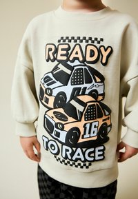 Hanorac crem cu un grafic mare cu două mașini de curse, una portocalie și una albastră, și textul „READY TO RACE” scris cu litere negre îndrăznețe.