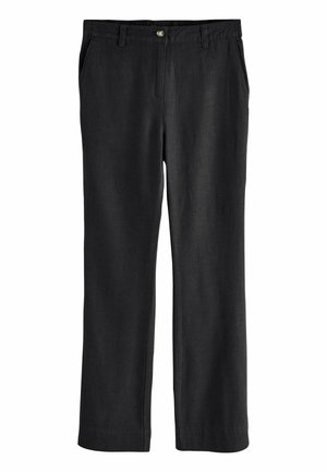 Femeie cu păr creț purtând o vestă neagră fără mâneci cu nasturi, pantaloni evazați negri și pantofi închiși la culoare cu buline albe.