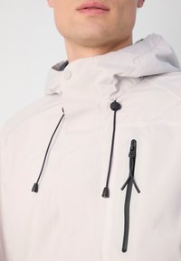 Light beige waterproof jacket with a hood, features black drawstrings and zip pocket. Smooth texture with matte finish.