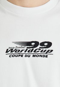 T-shirt bianca in cotone con una grafica nera stampata che recita "99 WorldCup Coupe du Monde." Collo rotondo e maniche corte.