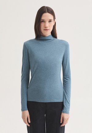 Frau mit glatten braunen Haaren, die einen blauen Langarmrollkragen und schwarze Hose trägt, stehend vor einem einfarbigen hellen Hintergrund.