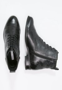 Černé kožené kotníkové boty mají hladký povrch, šněrování vpředu, boční zip a brogue detaily podél švů a špičky.