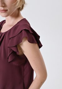 Frau trägt eine ärmellose dunkelviolette Bluse mit geschichteten Rüschendetails an den Schultern vor einem schlichten Hintergrund.