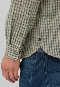 Beige en groene geruite shirtmouw met knopen, gedragen over blauwe denim jeans met een zichtbare hand die naast de heup rust.