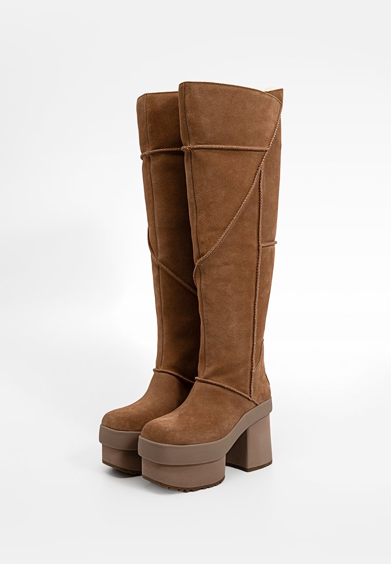 Bottes marron en daim jusqu'au genou avec une épaisse semelle plateforme et un talon block, présentant un design de couture unique et une texture lisse.
