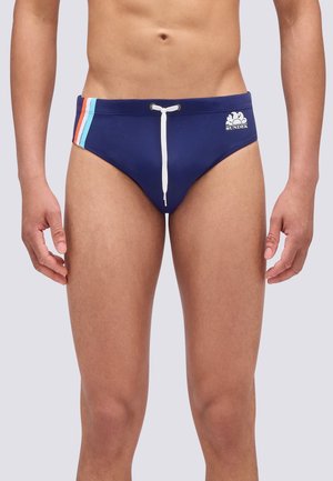 Uomo che indossa slip da bagno blu navy con coulisse bianca e strisce rosse, bianche e blu sull'anca sinistra, in piedi davanti a uno sfondo semplice.
