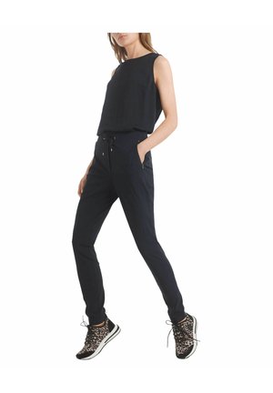 Femme portant un haut noir sans manches, un pantalon noir fuselé avec une taille à cordon, et des baskets à imprimé léopard, debout avec une main dans la poche.