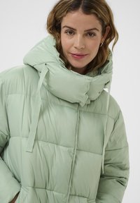 Kobieta o jasnobrązowych włosach, ubrana w jasną zieloną kurtkę puchową z wysokim kołnierzem i kapturem, delikatnie się uśmiechająca na jednolitym tle.