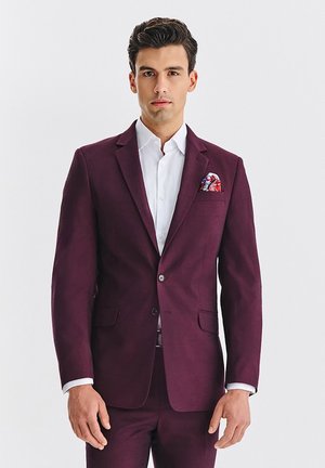 Burgundowa marynarka z klapą z wycięciem i dwoma przednimi kieszeniami. Noszona na białej koszuli z wzorzystą poszetką włożoną do kieszeni.