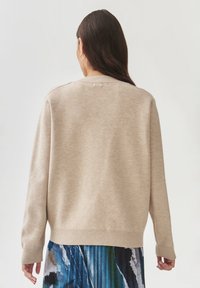 Beige gebreide trui met lange mouwen en geribde manchetten, met een ronde hals en een accentdetail op de rug. Soepele textuur en losse pasvorm.