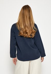 Tumši zils garām piedurknēm krekls ar brīvu piegriezumu, pogu detaļas uz piedurknēm un gludu tekstūru; valkāts virs gaišām biksēm.