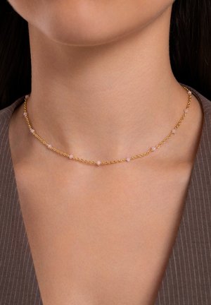 Gouden ketting met kleine roze kralen, met een verfijnd ontwerp en fijne textuur, rustend op een blootgestelde nek tegen een bruine achtergrond.