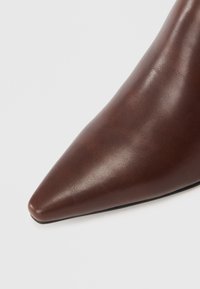Acercamiento de un zapato de cuero marrón de punta afilada con textura suave y un ligero brillo sobre un fondo blanco.