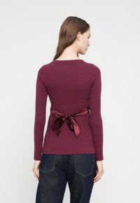 Μπλούζα μπορντό με μακριά μανίκια, εφαρμοστή μέση και λεπτομέρεια σατέν φιόγκου στο πίσω μέρος, συνδυασμένη με σκουρόχρωμο μπλε τζιν παντελόνι. Λείο ύφασμα, απλός σχεδιασμός.