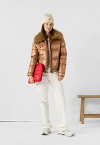 Giacca a vento in bronzo metallico con colletto in pelliccia sintetica, top crema, pantaloni bianchi e sneakers argento. Tiene una borsa rossa trapuntata.