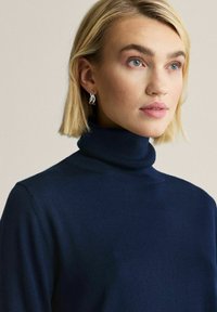 Vaaleatukkainen sinisilmäinen nainen, jolla on hopeiset korvakorut ja tummansininen pooloneule, vaaleaa neutraalia taustaa vasten.