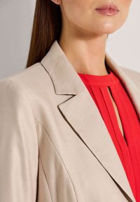 Beige op maat gemaakte blazer met een geknipte revers, gecombineerd met een rode geplooide top met een sleutelgatdetail bij de halslijn. Gladde texturen zijn zichtbaar.