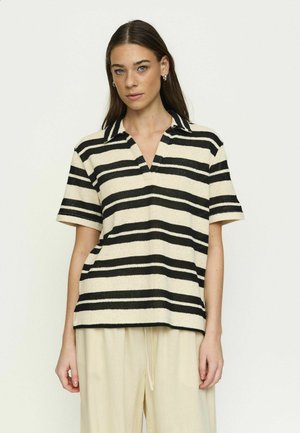 Woman wearing a short-sleeve black and beige horizontal striped polo shirt with beige loose pants against a plain background.