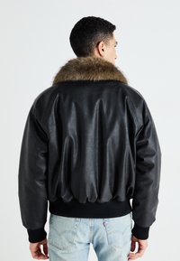 Giubbotto bomber in pelle nera con colletto in pelliccia marrone, polsini e vita a costine, e una vestibilità rilassata. Presenta una texture liscia e un retro arricciato.