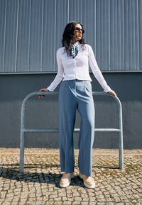 Femme portant un blouse texturée blanche, une écharpe bleue, un pantalon large bleu clair, des chaussures beiges et des lunettes de soleil, appuyée sur une rambarde en métal à l'extérieur.