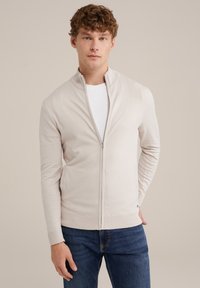 Hellbeige Zip-Up-Pullover mit geripptem Kragen und Bündchen, aus weichem Material. Kombiniert mit dunkelblauen Jeans.