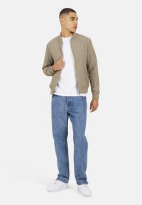 Beżowa, teksturowana kurtka bomberka z zamkiem z przodu, zestawiona z białą koszulką i niebieskimi dżinsami, dopełniona białymi sneakersami.