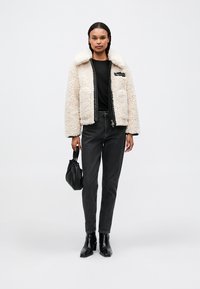 Hvid shearling-stil jakke med en sort lynlås, to frontlommer, båret over en sort top, parret med sorte jeans og ankelstøvler.