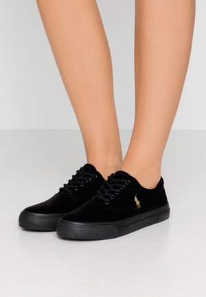 Czarne niskie trampki na płaskiej podeszwie z gumowymi podeszwami i małym złotym logo konia po boku, noszone na bose stopy.