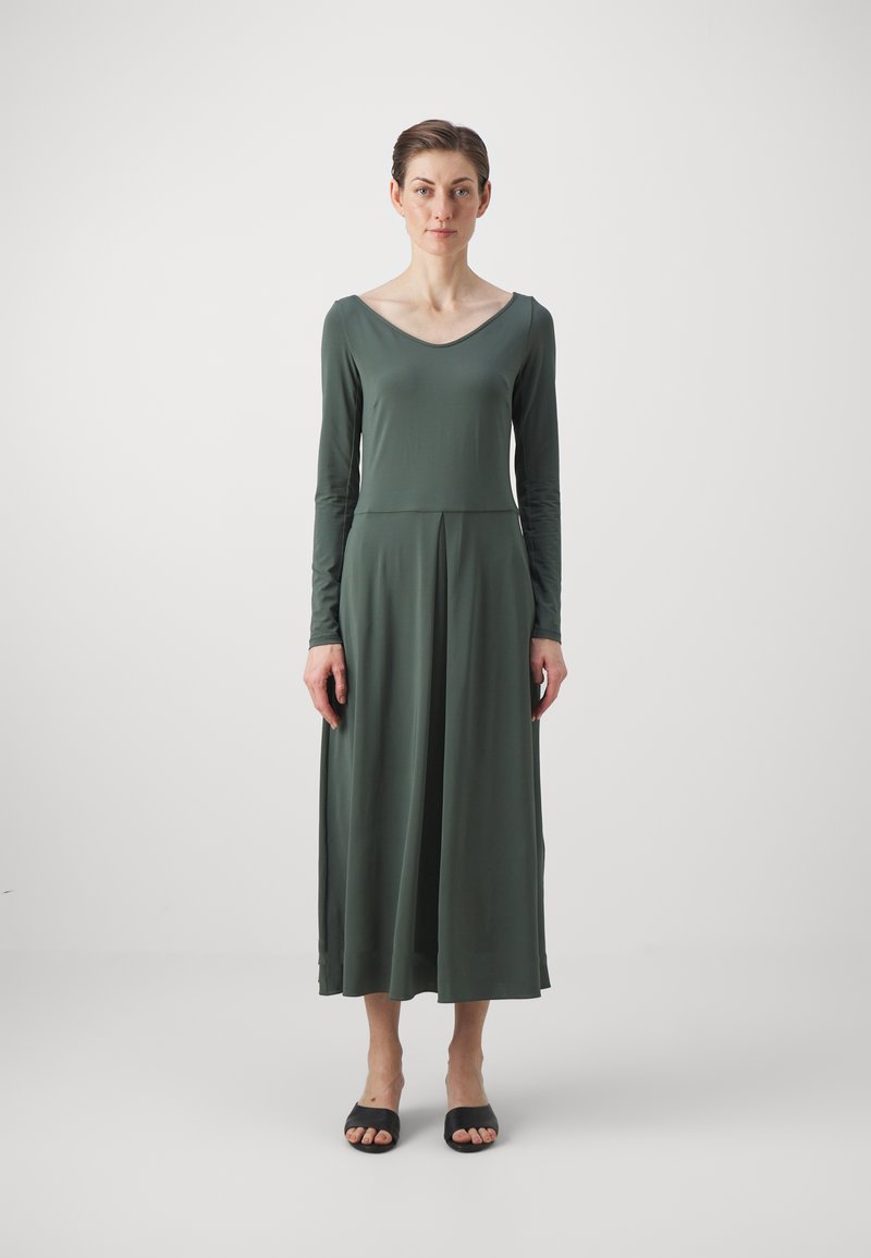 Robe à manches longues de couleur vert foncé en tissu lisse, avec un corsage ajusté et une jupe évasée plissée. Sandales noires à bout ouvert.